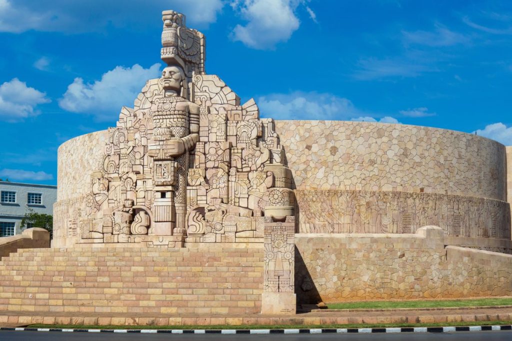Monument en pierre avec des sculptures complexes et un grand personnage central, entouré d'un mur de pierre rond et d'un ciel bleu, situé dans une rue de la ville.