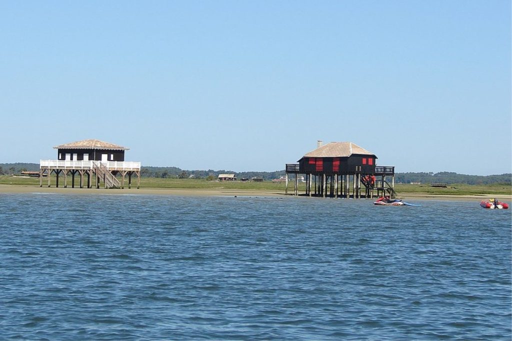 Deux maisons en bois sur pilotis se dressent près d'un rivage herbeux, avec une eau bleue calme au premier plan et un ciel clair au-dessus.