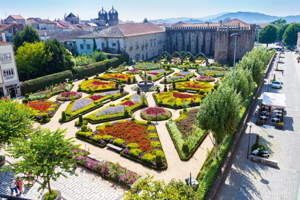 Vue aérienne d'un jardin à la française avec des parterres de fleurs colorés, des allées et une fontaine centrale, adjacent à un vieux bâtiment en pierre et entouré d'arbres et d'une rue.