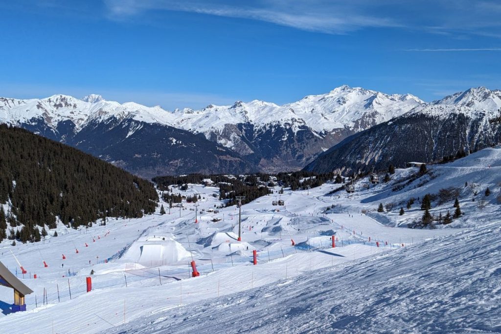 Une station de ski enneigée avec des pistes balisées, entourée de pins et de montagnes sous un ciel bleu clair.