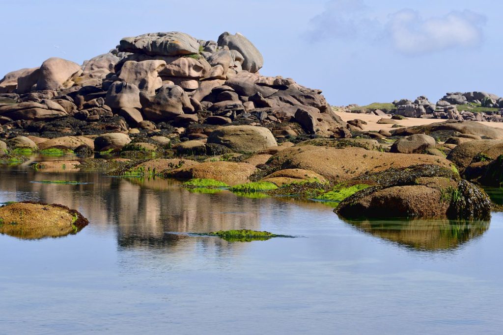De gros rochers lisses et des roches couvertes de taches d'algues vertes se reflètent dans des bassins de marée calmes sous un ciel clair.