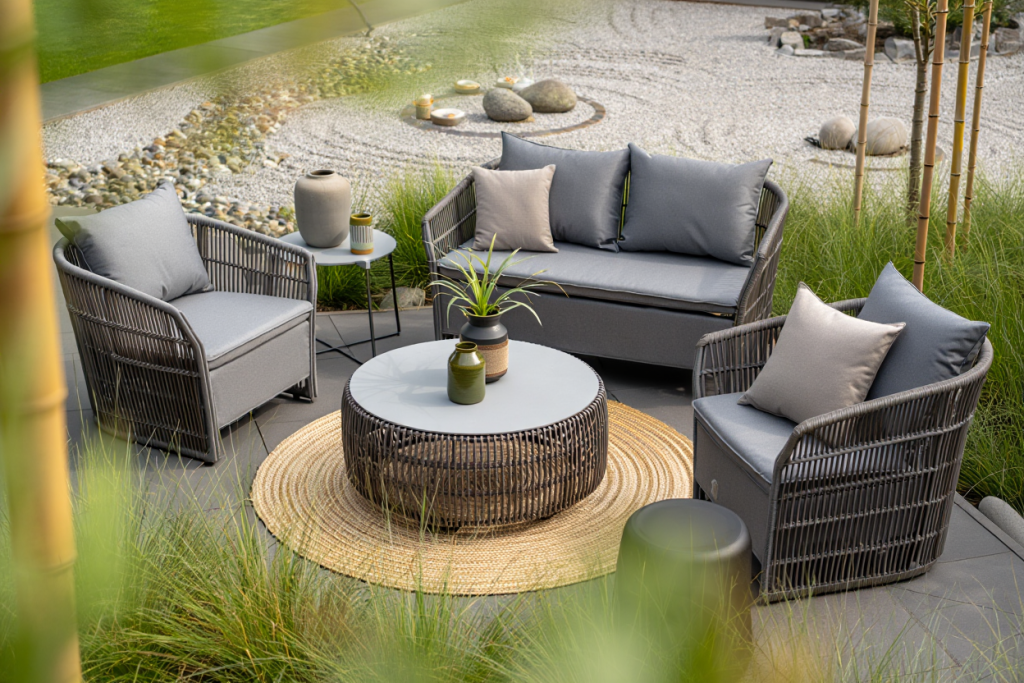 Terrasse moderne avec des meubles en osier, des coussins gris, une table basse ronde, des plantes en pot et un tapis tressé, le tout dans un jardin paysager avec du gravier et des rochers.