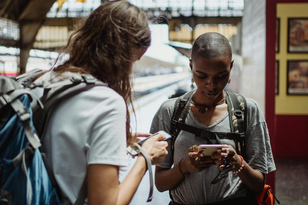 Deux personnes portant des sacs à dos se tiennent à l'intérieur de ce qui semble être une gare ou un lieu de transit, regardant vers le bas et utilisant leurs smartphones.