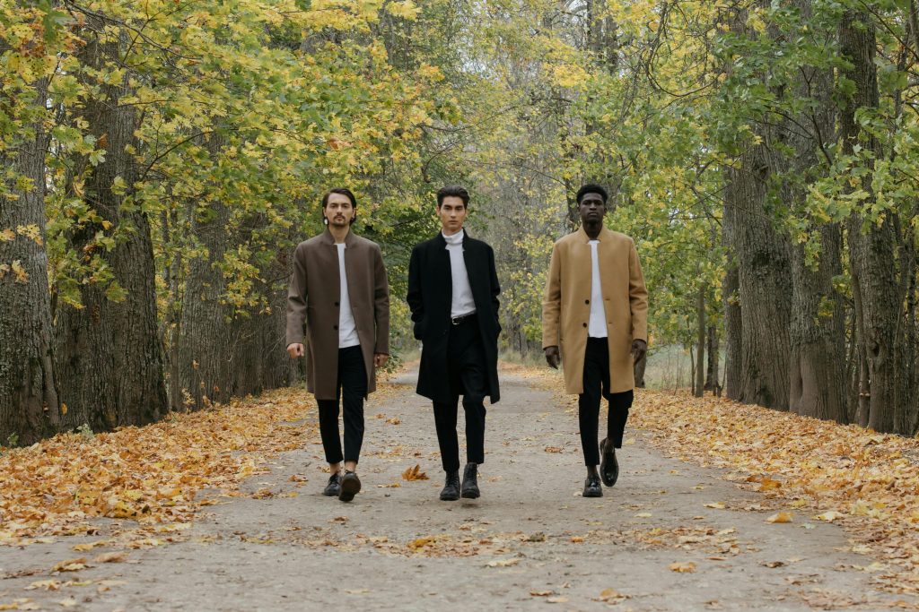 Trois hommes vêtus de manteaux élégants marchent sur un chemin bordé d'arbres couverts de feuilles d'automne.