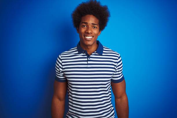 Un homme à la coiffure afro sourit à l'appareil photo, vêtu d'un polo rayé bleu et blanc, sur un fond bleu uni.