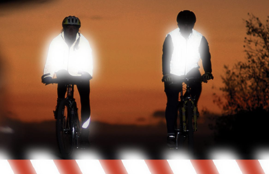 Deux cyclistes roulent au crépuscule ; l'un porte un gilet réfléchissant qui brille fortement, tandis que l'autre porte un gilet non réfléchissant, qui apparaît plus sombre sur le fond du coucher de soleil.