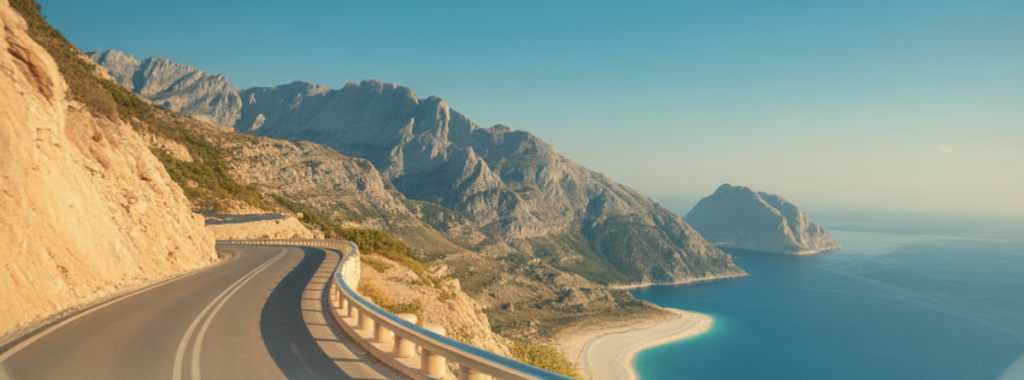 Une route côtière sinueuse longe des falaises rocheuses et offre une vue sur les montagnes et la mer bleue sous un ciel dégagé.