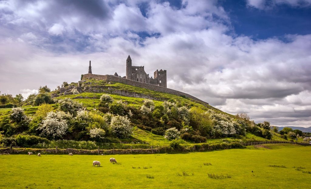 Un ancien château de pierre se dresse au sommet d'une colline herbeuse, entourée d'arbres et de verdure, tandis que des moutons paissent dans le champ en contrebas, sous un ciel partiellement nuageux.