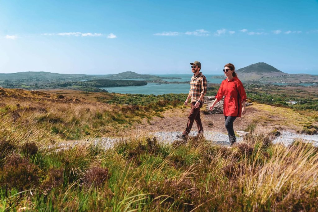 Deux personnes marchent sur un chemin de gravier à travers des collines herbeuses avec un lac et des montagnes en arrière-plan sous un ciel bleu clair.