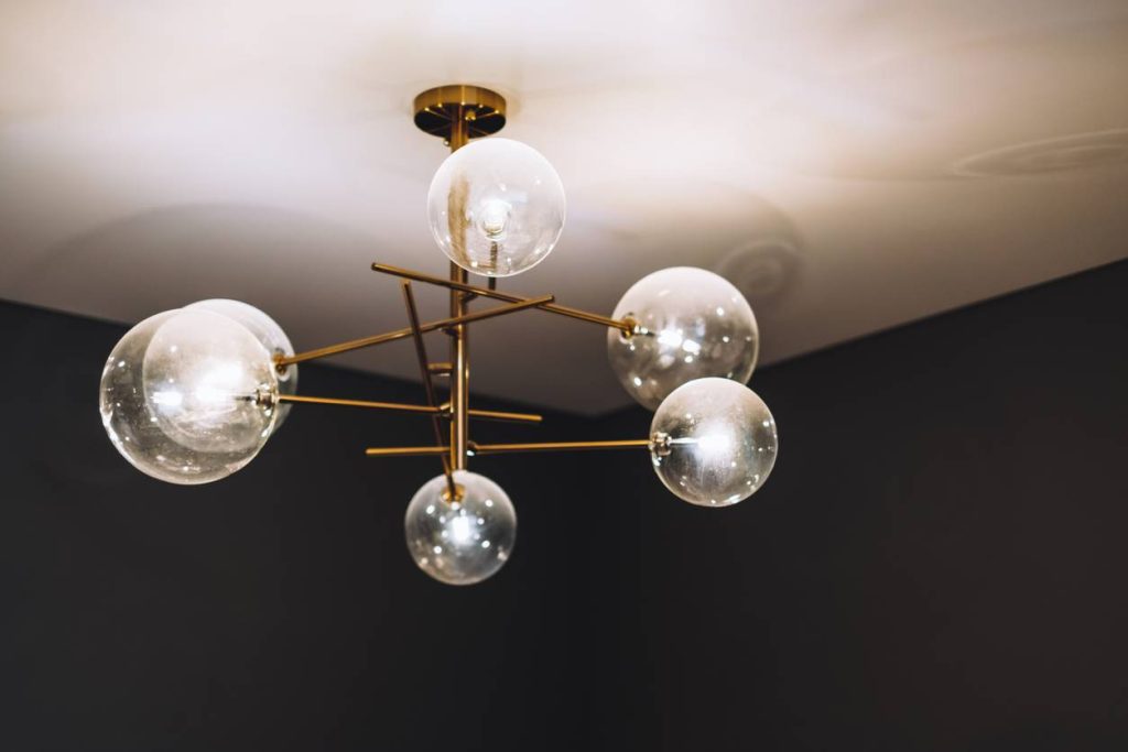 Plafonnier moderne avec un cadre en métal doré et six ampoules globe en verre transparent, monté sur un plafond blanc dans une pièce aux murs sombres.