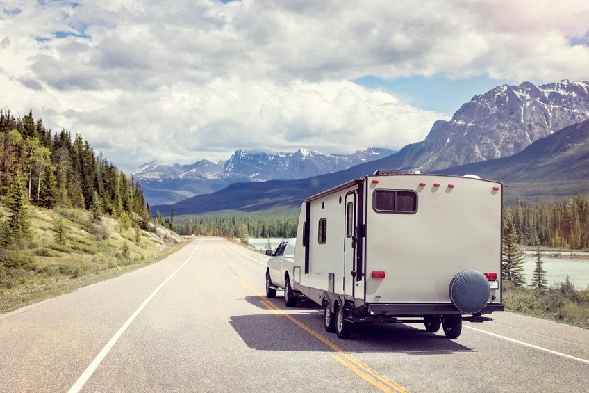 Une camionnette blanche tirant une caravane roule sur une route de montagne pittoresque le long d'une rivière, entourée d'arbres et de sommets enneigés sous un ciel partiellement nuageux.