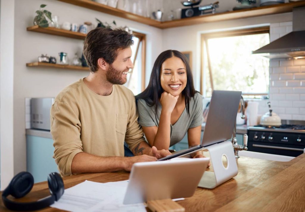 Un homme et une femme sont assis à une table de cuisine, utilisant un ordinateur portable et une tablette, souriant et discutant, avec des papiers et des écouteurs sur la table autour d'eux.