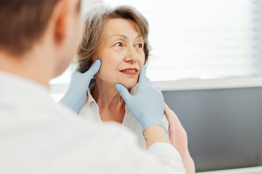 Un médecin portant des gants bleus examine le visage d'une femme âgée lors d'une consultation médicale dans une pièce lumineuse.