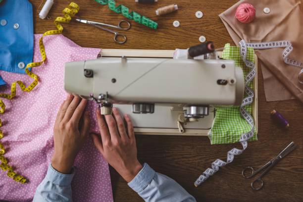 Personne cousant du tissu à pois rose sur une machine à coudre, entourée de tissus colorés, de rubans à mesurer et d'outils de couture sur une table en bois.