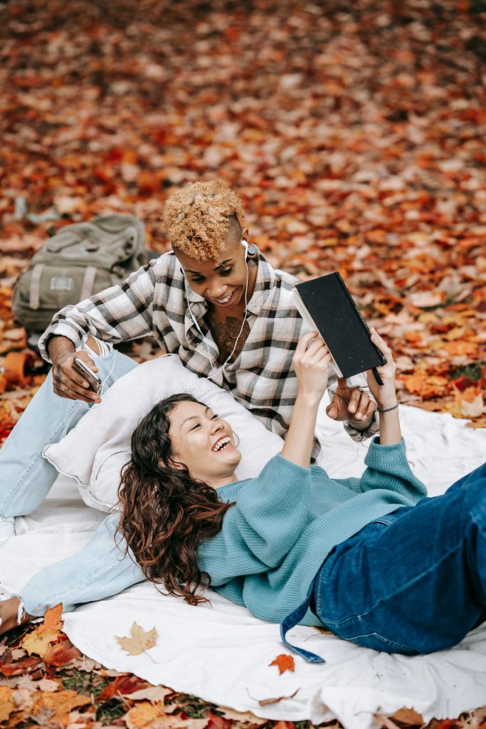 Deux personnes se détendent sur une couverture au milieu de feuilles d'automne, partageant des écouteurs et regardant ensemble une tablette. L'un d'eux tient une tasse de café et un sac à dos est posé sur le sol à proximité.