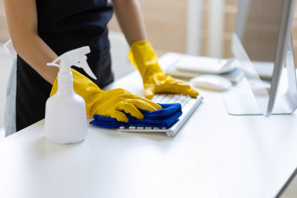 Une personne portant des gants jaunes nettoie un clavier d'ordinateur avec un chiffon bleu sur un bureau blanc ; un flacon pulvérisateur et un moniteur se trouvent à proximité.