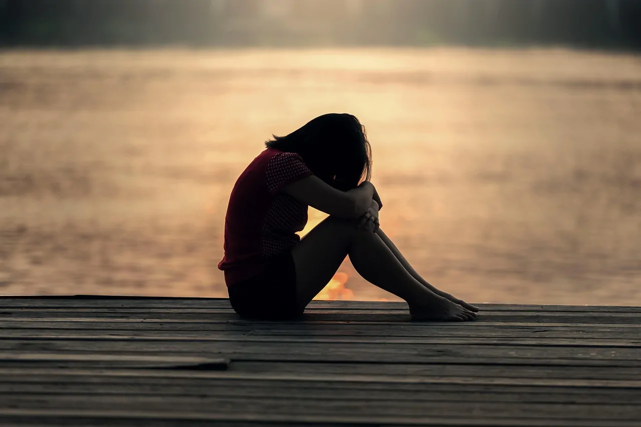 Une personne est assise seule sur un ponton en bois au bord de l'eau, au coucher du soleil, les genoux repliés et la tête posée sur ses bras.