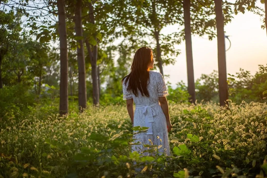 Une femme vêtue d'une robe blanche se tient debout dans un champ baigné de soleil, entourée de hautes herbes et d'arbres, le regard tourné vers la lumière.