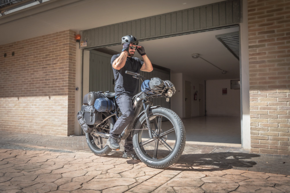 Une personne vêtue de noir et portant un casque ajuste ses lunettes de soleil, assise sur un vélo électrique chargé, devant un garage.