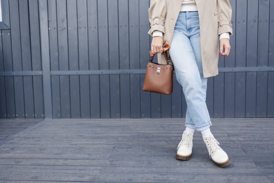 Personne portant un trench beige, un jean bleu clair et des bottes blanches, tenant un sac à main marron, debout devant une clôture en bois foncé.