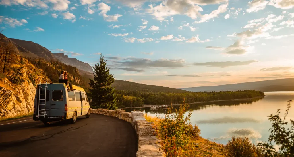 Une camionnette garée sur une route de montagne pittoresque au bord d'un lac au lever du soleil, avec les montagnes, les arbres et un ciel partiellement nuageux qui se reflètent dans l'eau.