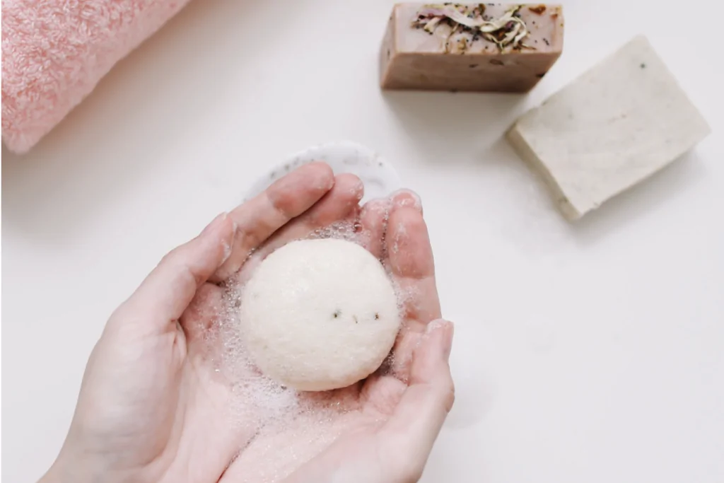 Des mains tenant une bombe de bain ronde et savonneuse, recouverte de mousse, à côté de serviettes roses et blanches et de deux pains de savon rectangulaires posés sur une surface blanche.