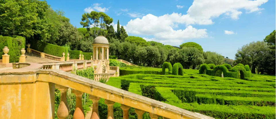 Un jardin à la française avec des haies soigneusement taillées, une balustrade en pierre et un petit pavillon surmonté d'un dôme, sous un ciel bleu parsemé de nuages.