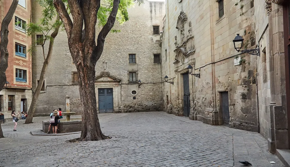 Une place pavée où se dresse un grand arbre, un vieux bâtiment en pierre aux portes richement décorées, et des gens rassemblés près d'une petite fontaine.