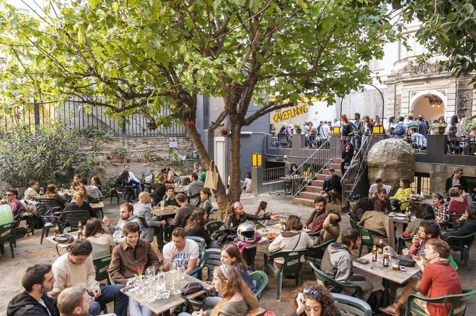 Une terrasse de café en plein air où de nombreuses personnes sont assises à des tables sous les arbres, en train de manger, de boire et de discuter. D'autres personnes sont rassemblées sur une terrasse surélevée à l'arrière-plan.
