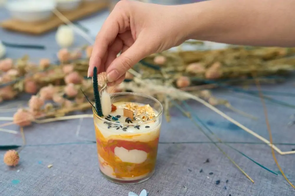 Une main tient un brin d'herbe séchée au-dessus d'un verre rempli d'une boisson laiteuse à plusieurs couches, garnie de graines et d'avoine, posé sur une table ornée de fleurs séchées.