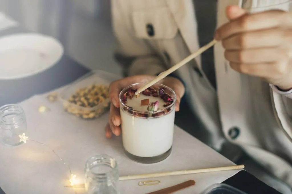 Une personne remue une bougie blanche dans un bocal en verre surmonté de fleurs séchées, à l'aide d'un bâtonnet en bois. Du matériel de loisirs créatifs et des bocaux sont posés sur la table.