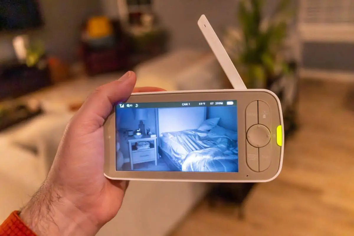 Une main tient un babyphone qui diffuse une vidéo en direct montrant un lit vide, soigneusement fait, à côté d'une table de chevet dans une chambre à la lumière tamisée.