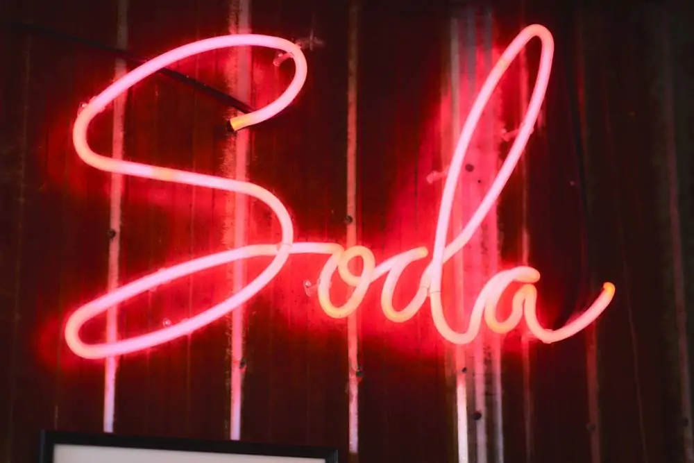 Une enseigne au néon affiche le mot « Soda » en lettres cursives sur un mur sombre strié de lignes verticales.