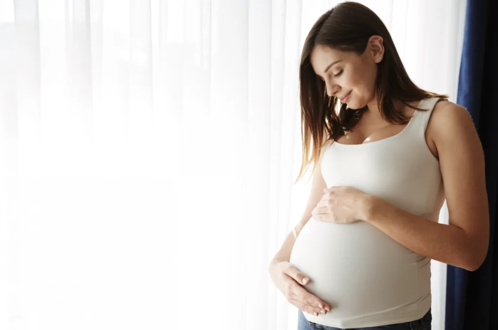 Une femme enceinte vêtue d'un débardeur blanc se tient près d'une fenêtre baignée de lumière, les deux mains posées délicatement sur son ventre.