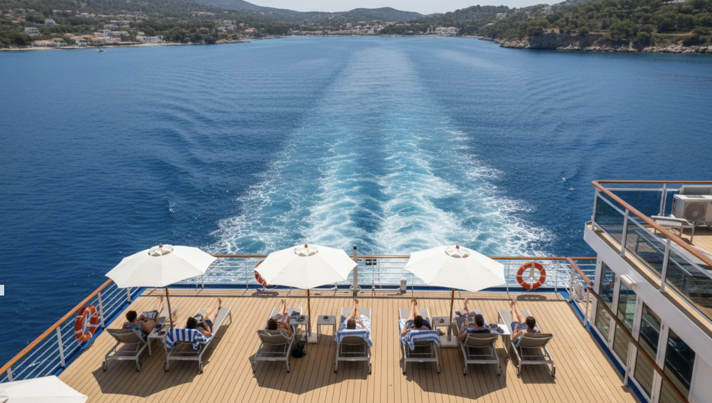 Des personnes sont allongées sur des transats à l'ombre de parasols à l'arrière d'un bateau de croisière, contemplant le sillage laissé par l'embarcation sur une mer bleue et calme, près d'un paysage côtier.