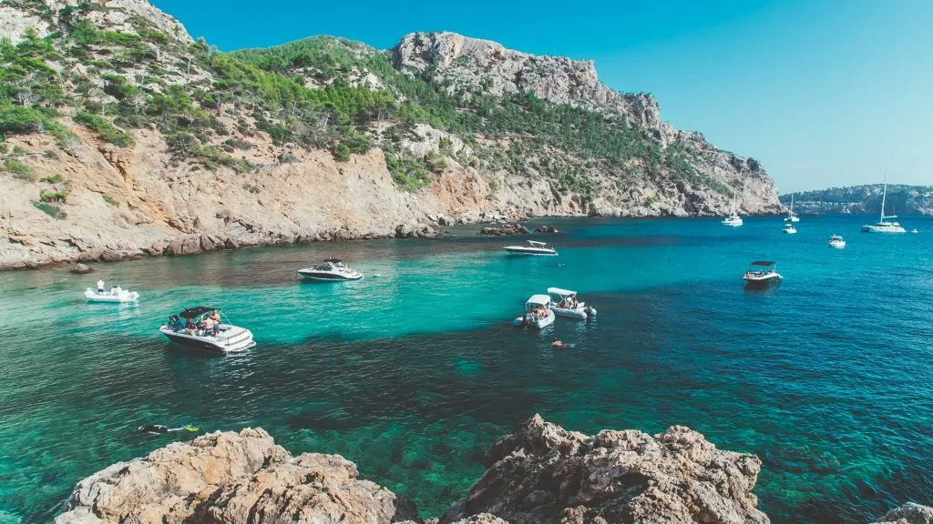 Des bateaux flottent sur une eau bleue et limpide dans une baie rocheuse, entourée de collines verdoyantes et de falaises, sous un ciel clair et radieux.