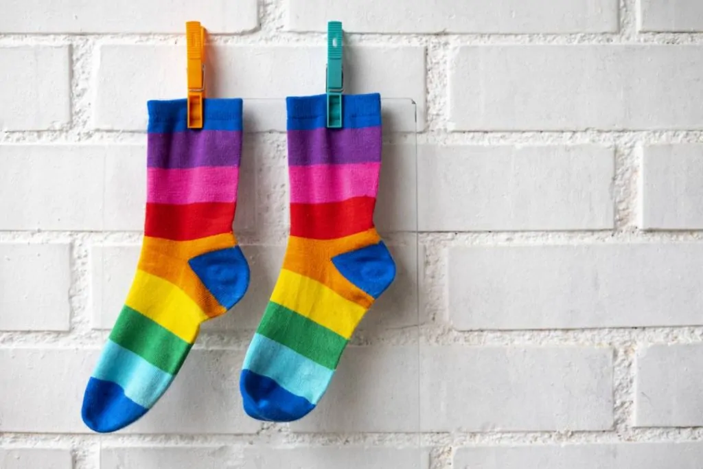 Deux chaussettes aux rayures arc-en-ciel sont accrochées à un mur de briques blanches à l'aide de pinces à linge orange et bleu sarcelle.