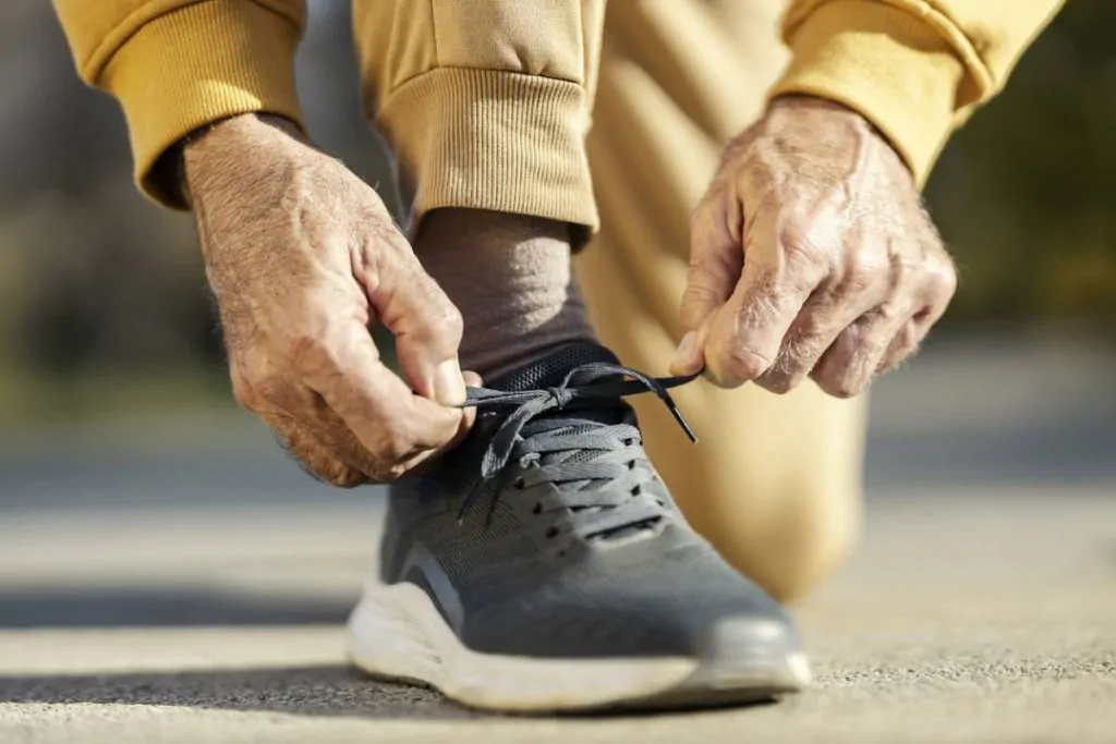 Gros plan sur les mains d'une personne âgée en train de lacer une chaussure de sport foncée, alors qu'elle porte un pantalon marron clair à l'extérieur.