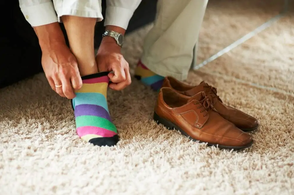 Une personne en train d'enfiler une chaussette rayée aux couleurs vives, assise sur un sol recouvert de moquette, avec des chaussures en cuir marron posées à proximité.