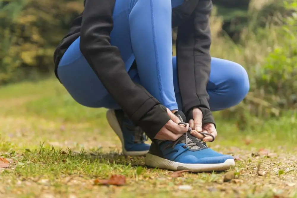 Une personne vêtue d'un legging bleu et d'une veste noire, en train de lacer ses chaussures de course bleues sur un chemin de terre à l'extérieur.